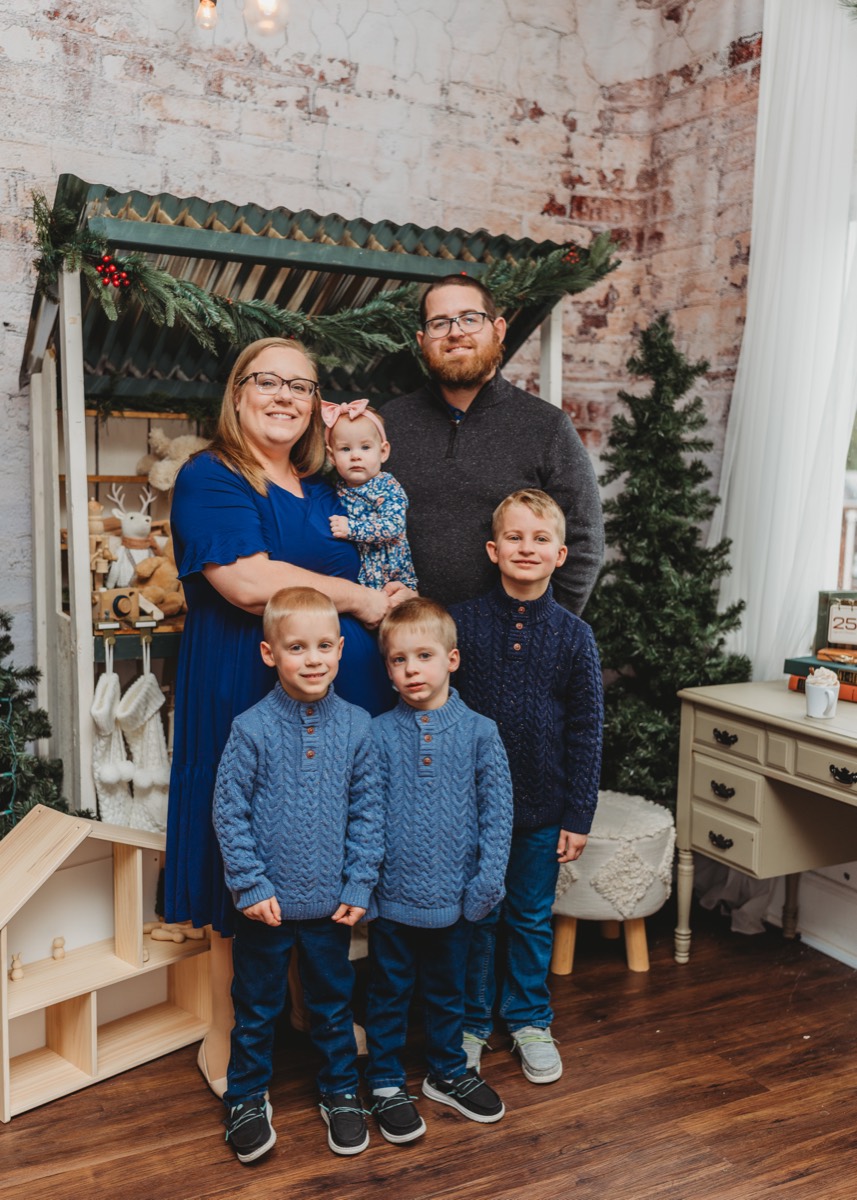 The Beville family in matching blue: Amanda holding Maci, Justin, and Luke, Tucker and Turner in cable-knit sweaters in front of a Christmas set.